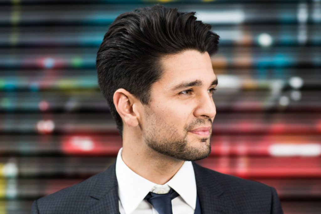Sam Dixon of Maddox Magic in a navy suit and tie, looking to one side against a blurred painted shutter, portrait by Mat Smith Photography, London