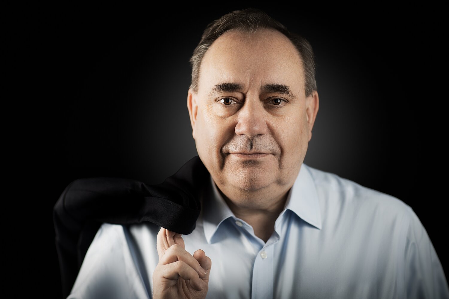 Alex Salmond, former First Minister of Scotland, in a pale blue shirt with his jacket over his shoulder, portrait on a black background