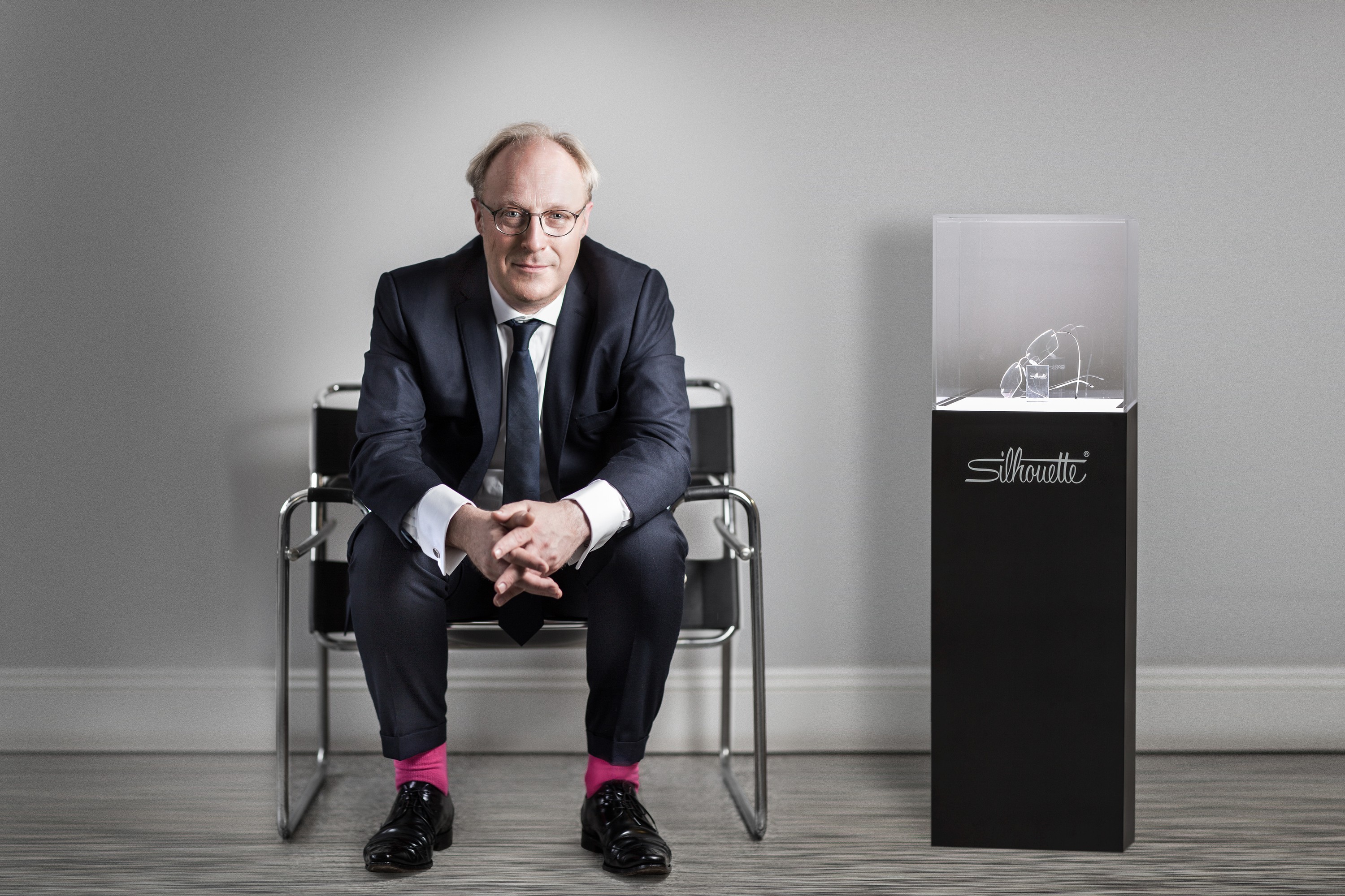 Company director in a navy suit and pink socks, seated on a chrome chair beside a display case of spectacles, portrait by Mat Smith Photography, Chiswick, London