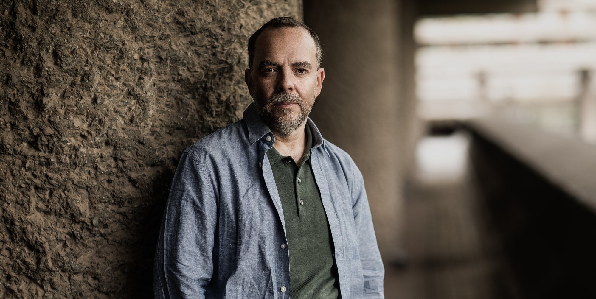 Matthew Reynolds in a blue linen shirt and green polo shirt, leaning against textured concrete at the Barbican, London